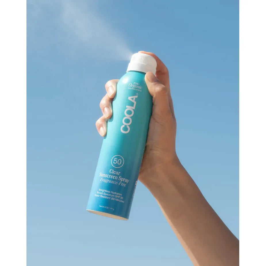 Hand holding a blue sunscreen spray can against a clear sky.