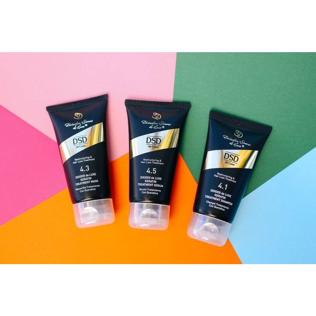 A travel set of DSD de Luxe hair care products including shampoo, hair mask, and hair serum, packaged in black tubes with gold and white accents, displayed in front of a  colorful background.